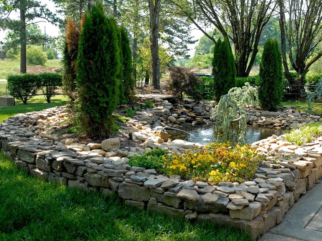A serene garden featuring a stone-bordered pond with lush greenery and flowering plants.