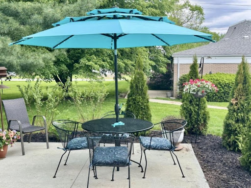 A tranquil outdoor patio setup featuring a teal umbrella over a table surrounded by chairs, set amid lush greenery.