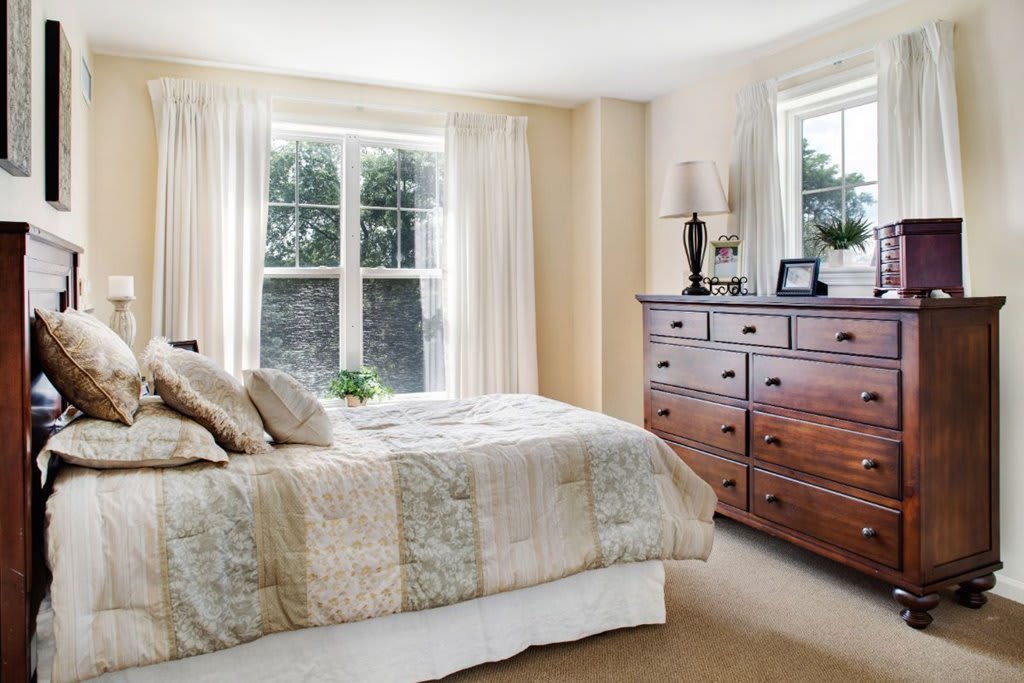 A cozy bedroom with a large bed and wooden dresser next to sunlit windows.