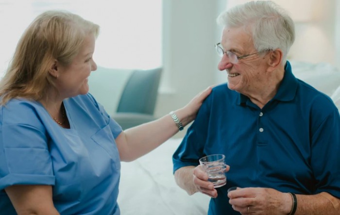 A nurse provides care and support to an elderly patient, who is holding a glass of water.