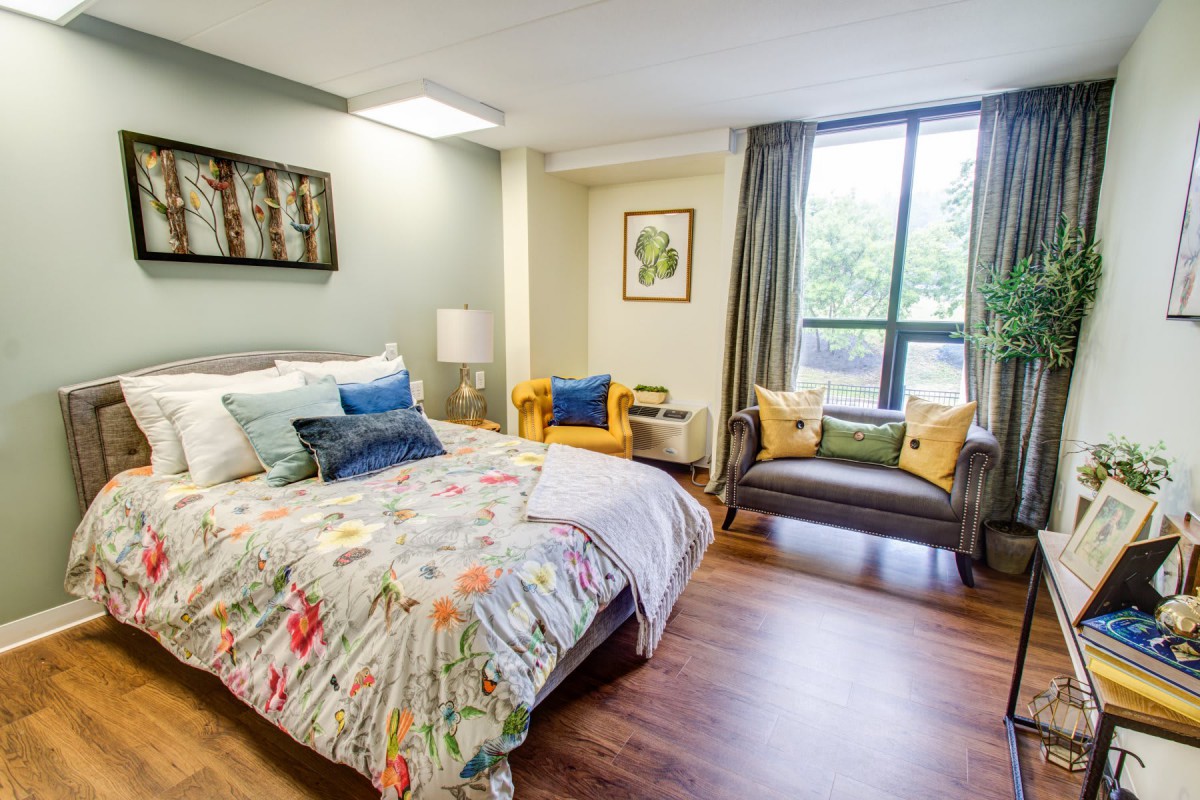 A modern bedroom with colorful bedding, a cozy armchair, and large windows allowing natural light.