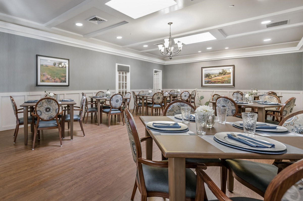 A well-lit dining room with neatly arranged tables and chairs, featuring elegant place settings and decorative artwork on the walls.