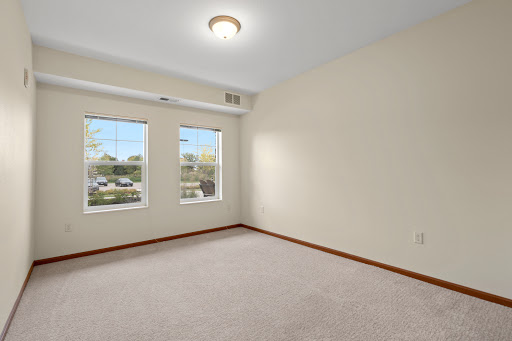 A spacious, empty room with two windows allowing natural light to flood in, featuring a carpeted floor and neutral wall colors.