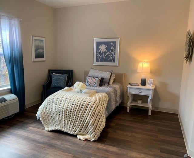 A cozy bedroom with a neatly made bed, a chair, and soft lighting from a lamp on the nightstand.