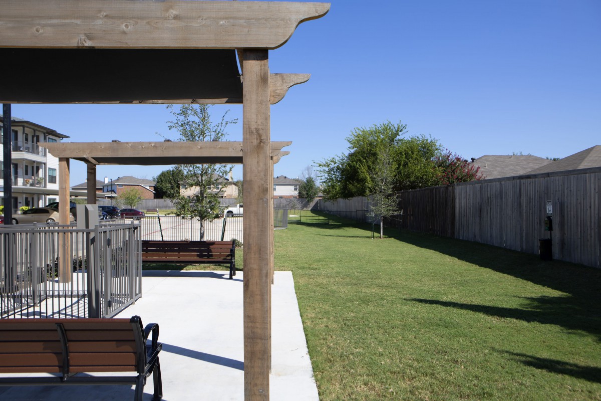 A sunny outdoor area featuring a pergola, benches, and a well-maintained lawn with trees.