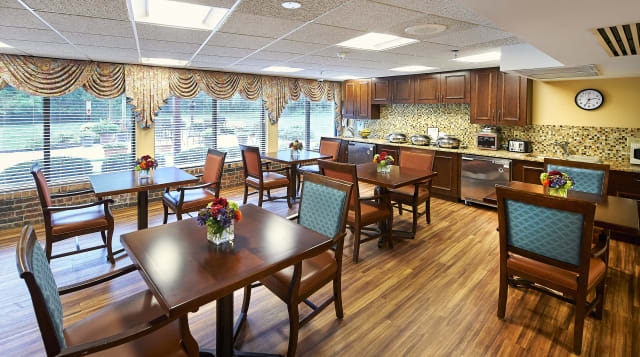 A cozy dining room with wooden tables and chairs, decorated with flowers and adjacent to a kitchen area.