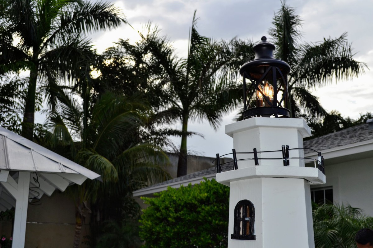 A small lighthouse with a glowing lamp stands against a backdrop of palm trees in the evening.