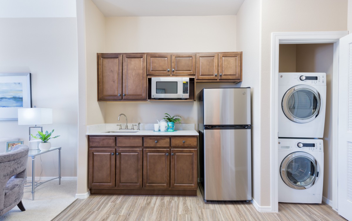 A compact kitchen area with wooden cabinets, a microwave, stainless steel refrigerator, and adjacent washer and dryer.