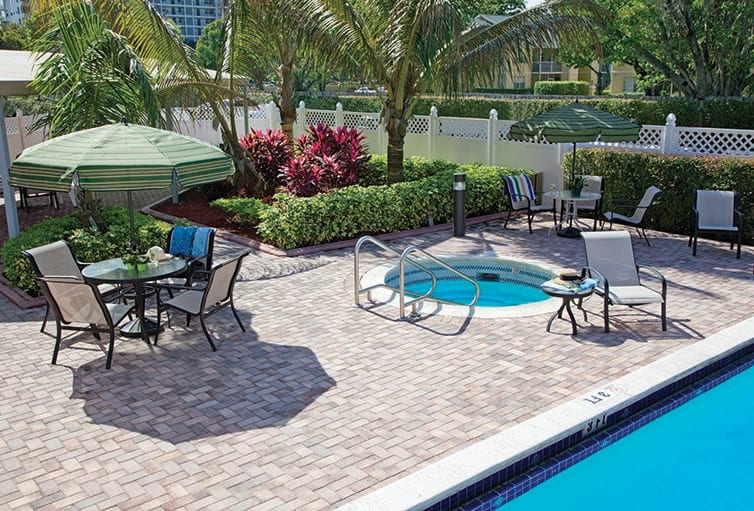 A sunny outdoor pool area with a hot tub, surrounded by patio furniture and tropical plants.