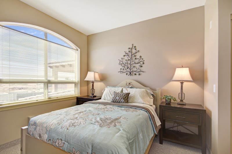 A cozy bedroom with natural light, featuring a bed with patterned bedding, two bedside lamps, and decorative wall art.