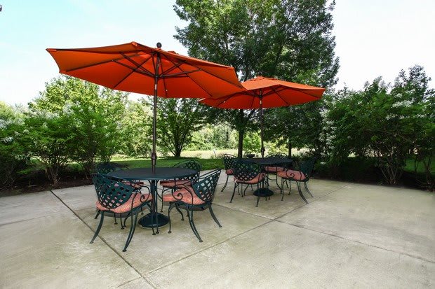 A well-furnished patio with tables and chairs under vibrant orange umbrellas, surrounded by lush greenery.