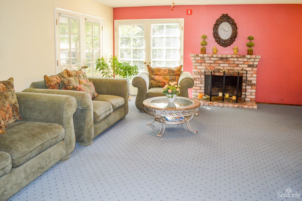 A cozy living room with a green sofa set, coffee table, and a brick fireplace, illuminated by natural light from large windows.