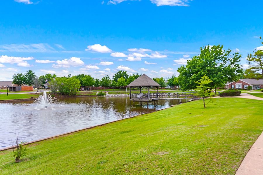 A peaceful park featuring a serene lake with a small pier, surrounded by green grass and trees under a clear blue sky.