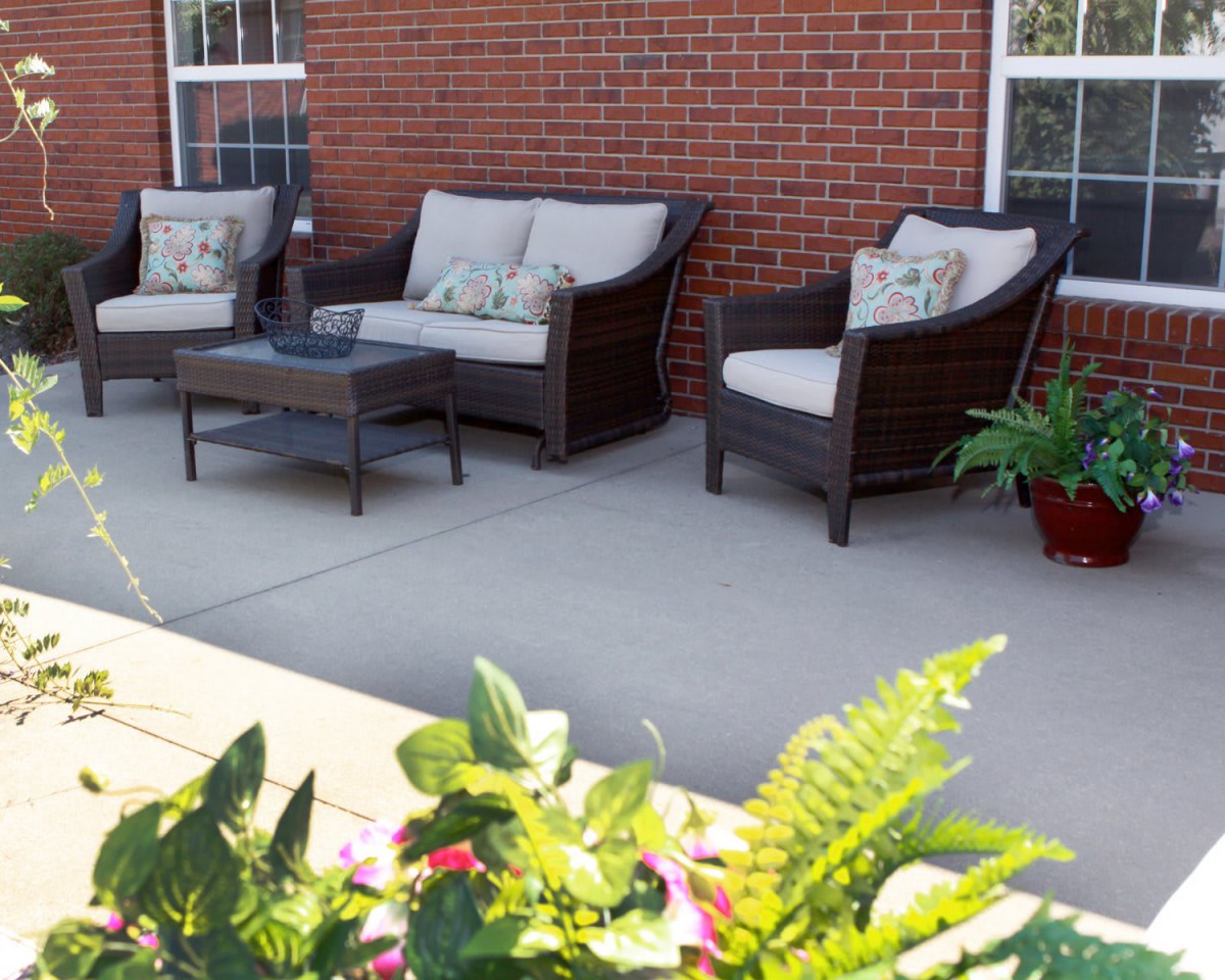 A cozy outdoor patio featuring wicker furniture, including two sofas and a chair, arranged around a coffee table with decorative plants nearby.