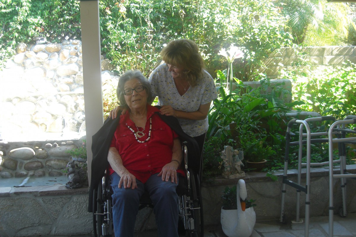 An elderly person in a wheelchair is being assisted by a caregiver in an outdoor garden setting.