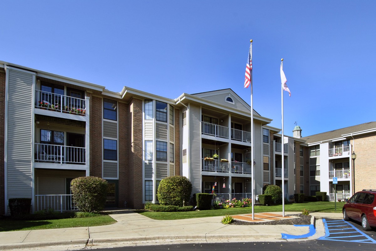 A three-story apartment building with balconies and flags flying in front, surrounded by well-maintained landscaping under a clear blue sky.