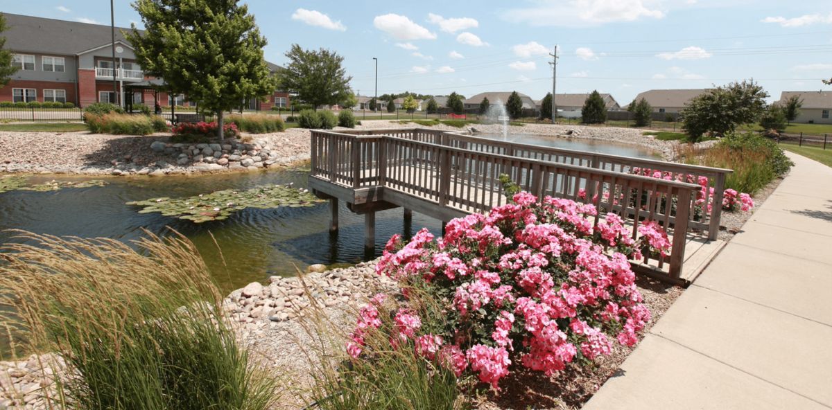 A scenic view of a garden with pink flowers beside a wooden bridge over a pond.