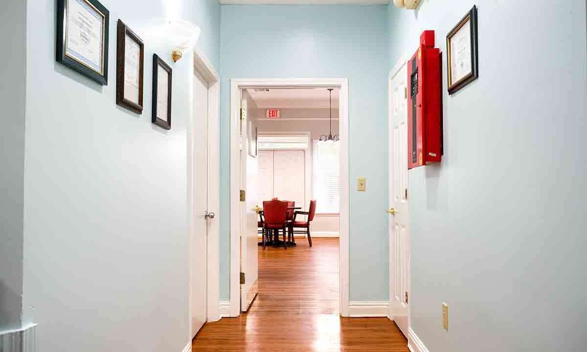 A brightly lit hallway with light blue walls and wooden flooring leading to a room with red chairs.