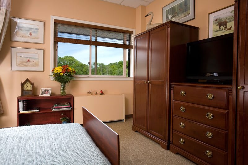 A cozy bedroom with wooden furniture, including a wardrobe and dresser, under natural light from a window.