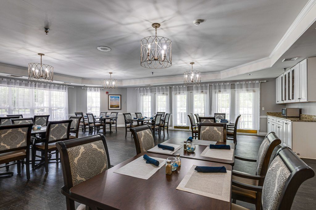 An elegant dining room with several tables set for guests, featuring chandeliers and natural lighting.