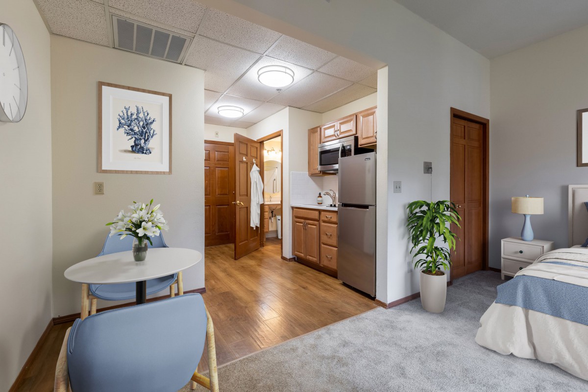 A modern apartment interior featuring a compact kitchen with wooden cabinetry and flooring, a small dining area, and decorative plants.