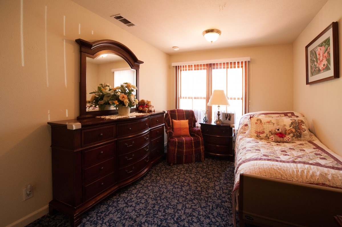 A cozy bedroom with wooden furniture, featuring a bed, dresser with mirror, and a lamp near the window.