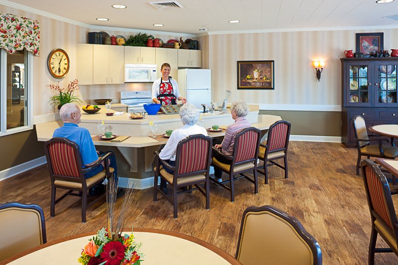 A group of elderly people sitting in a care home kitchen, interacting with a caregiver.