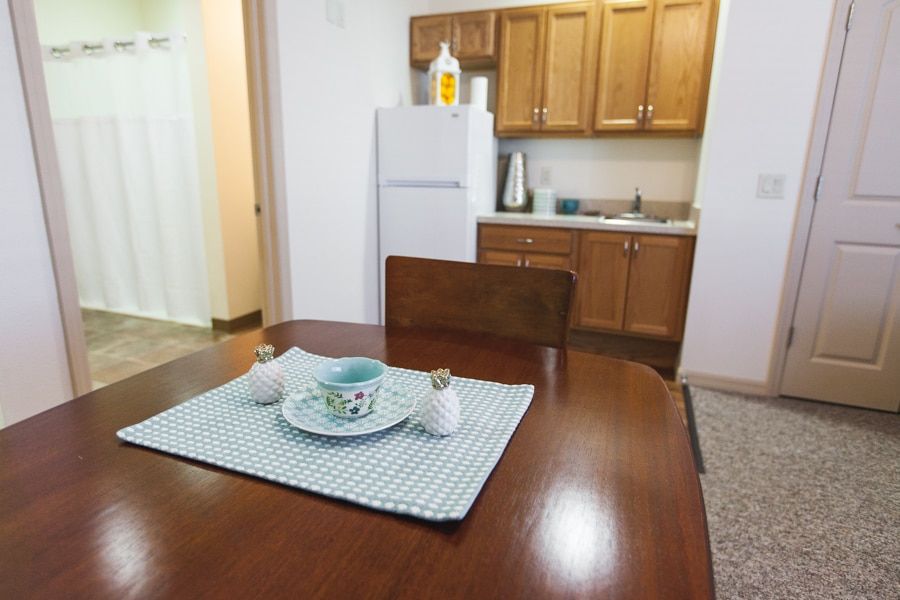 A cozy apartment kitchen featuring a wooden dining table set with a placemat and dishes, adjacent to cabinetry and a refrigerator.