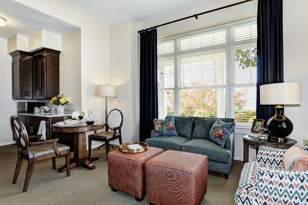 A cozy living and dining area with ample natural light, featuring a sofa, two ottomans, and a small dining table.
