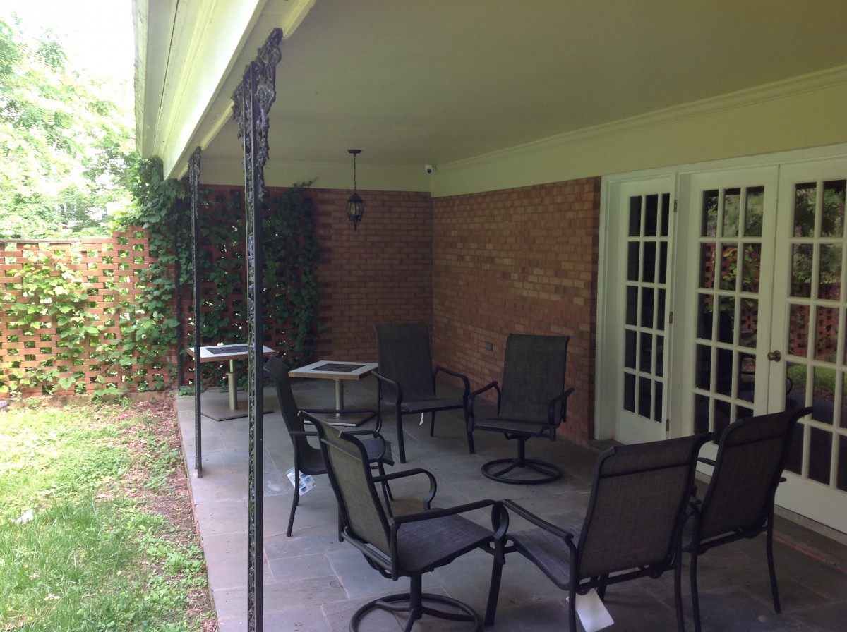 A cozy patio area with outdoor furniture set beneath a covered space, featuring a brick wall and surrounded by greenery.