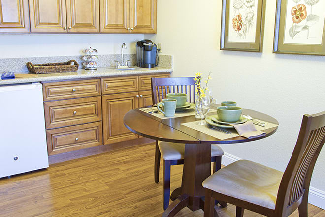 A cozy kitchen dining area featuring a wooden table set for two, surrounded by warm wooden cabinets.