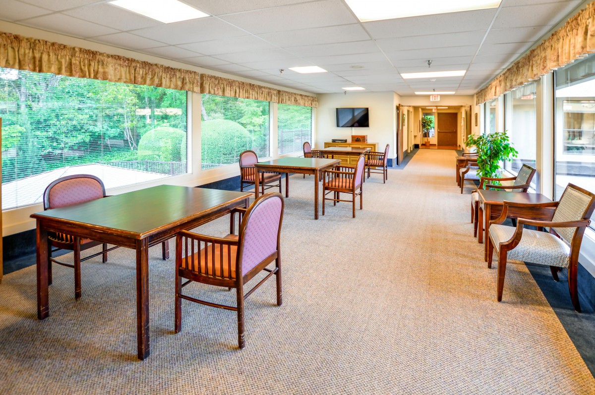 A bright dining area with tables and chairs, featuring large windows overlooking greenery.