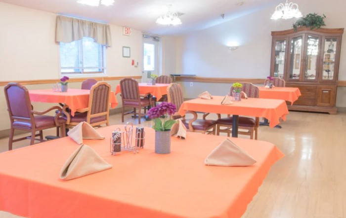A bright dining room with tables set for a meal, featuring orange tablecloths and floral centerpieces.