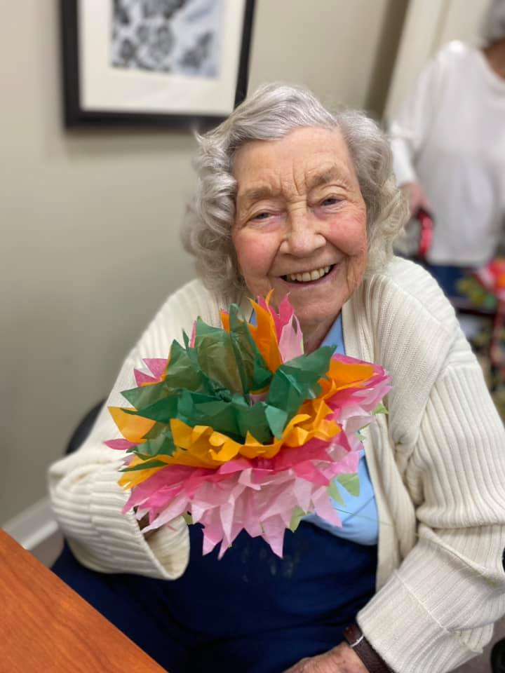 An elderly woman smiling and holding a colorful bouquet of flowers indoors.