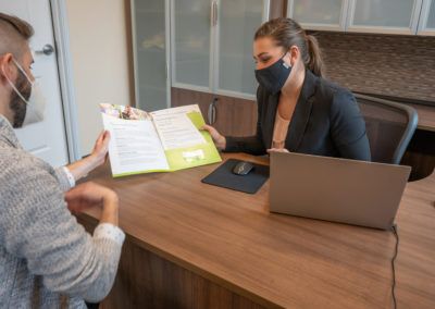 Two people in a business meeting with brochures and a laptop, wearing masks.