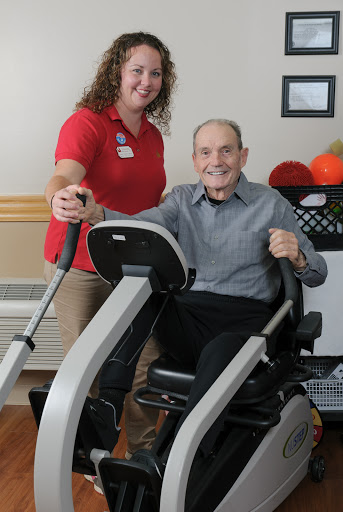 A caregiver assists an elderly man in exercising on a fitness machine, promoting health and rehabilitation.