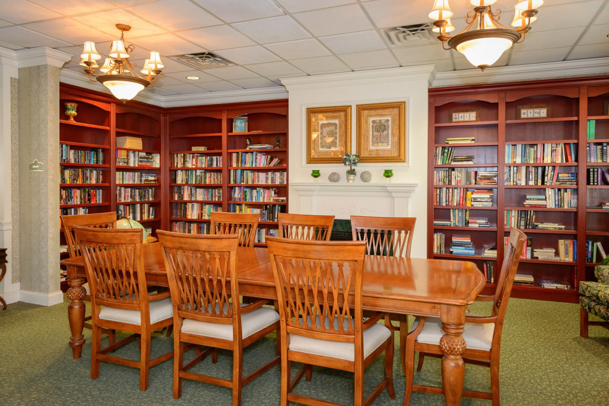 A cozy library setting with a large wooden table surrounded by chairs, flanked by bookshelves and illuminated by chandeliers.