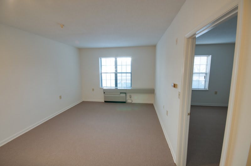 An empty apartment room with a large window and carpeted floor.