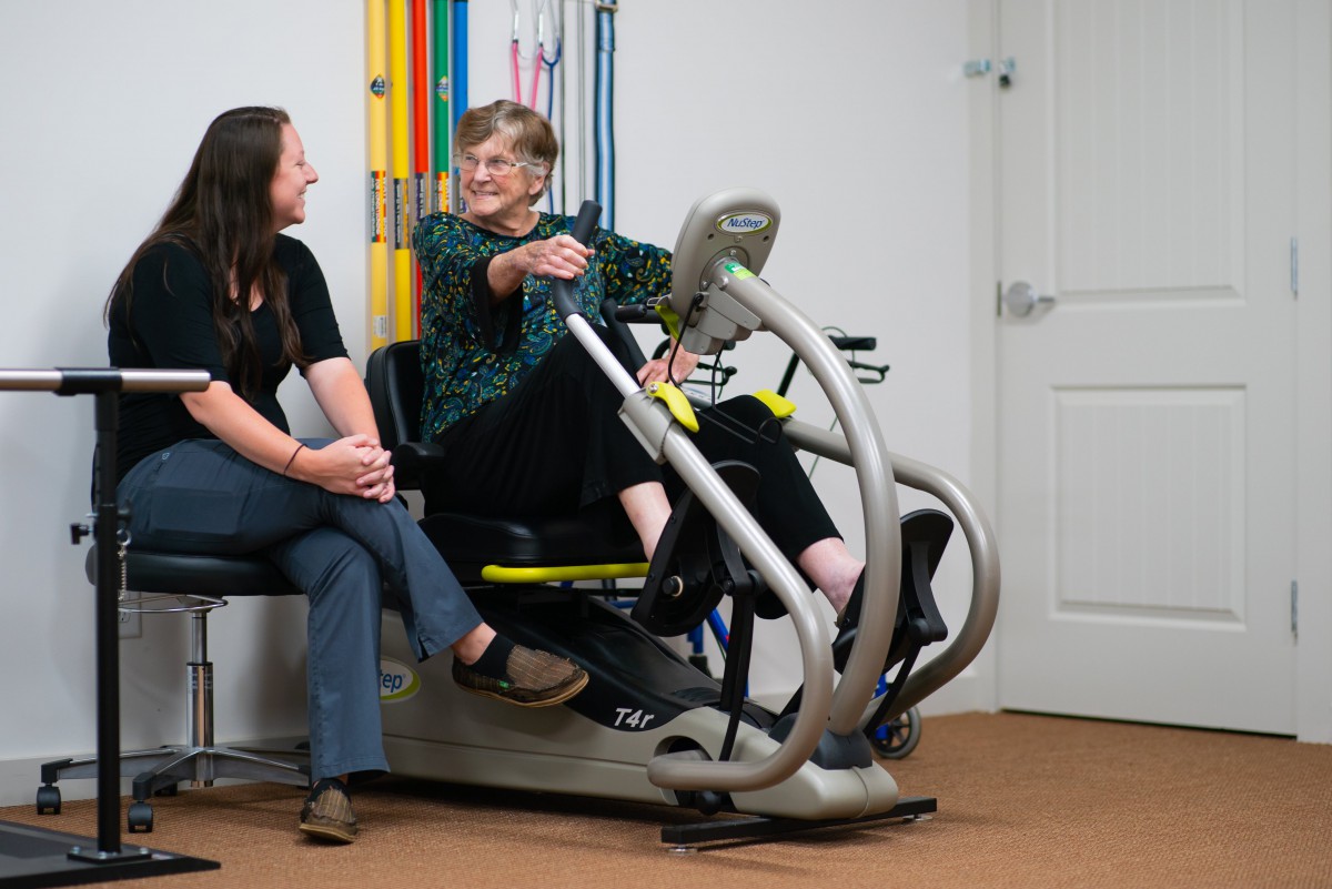 An elderly woman engages in a therapy session using a recumbent exercise bike, accompanied by a healthcare professional.