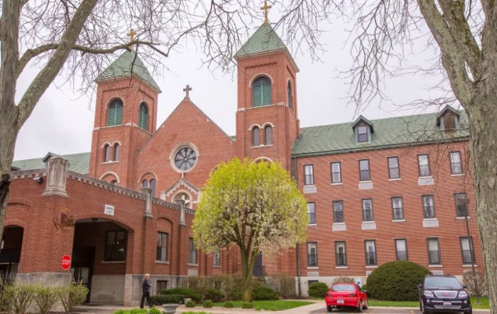 A brick church with twin towers and a green roof, surrounded by trees and a parked red car.