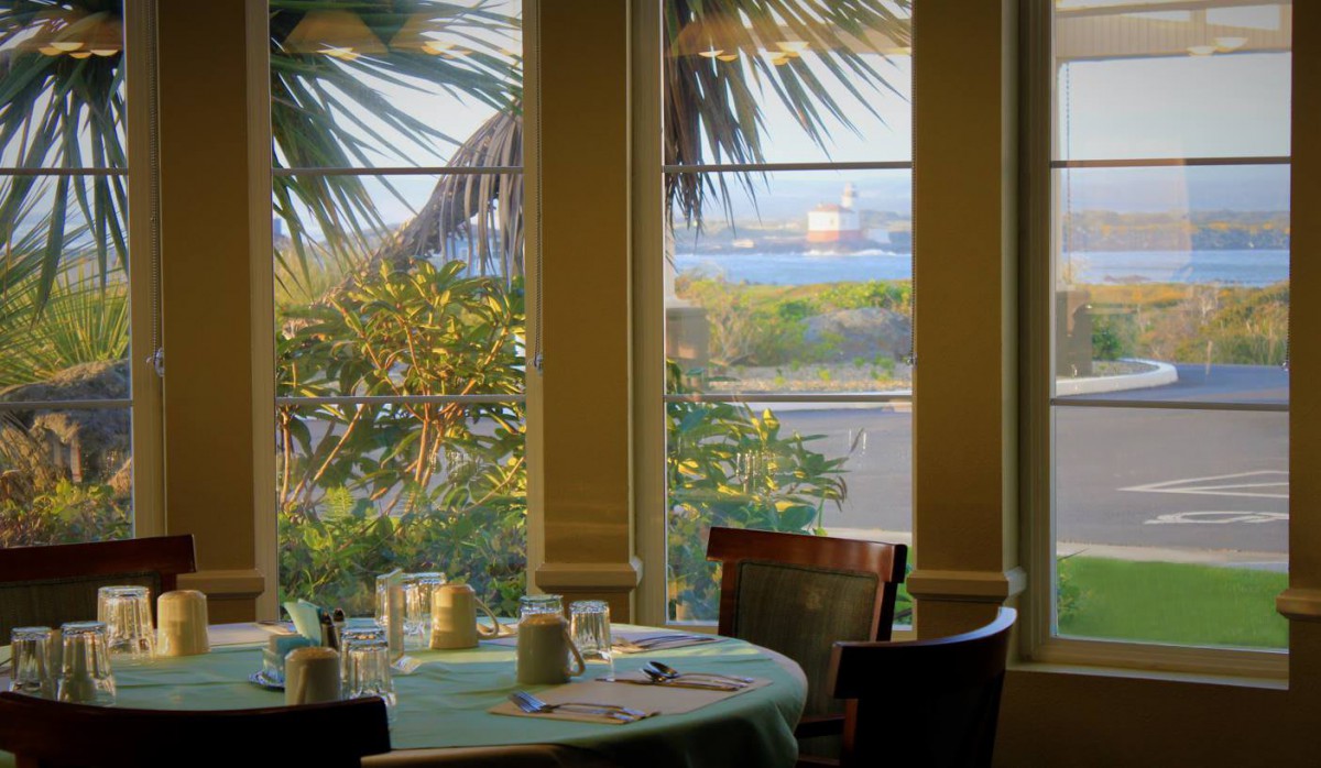 A peaceful restaurant dining area with a view of the ocean and palm trees through large windows.
