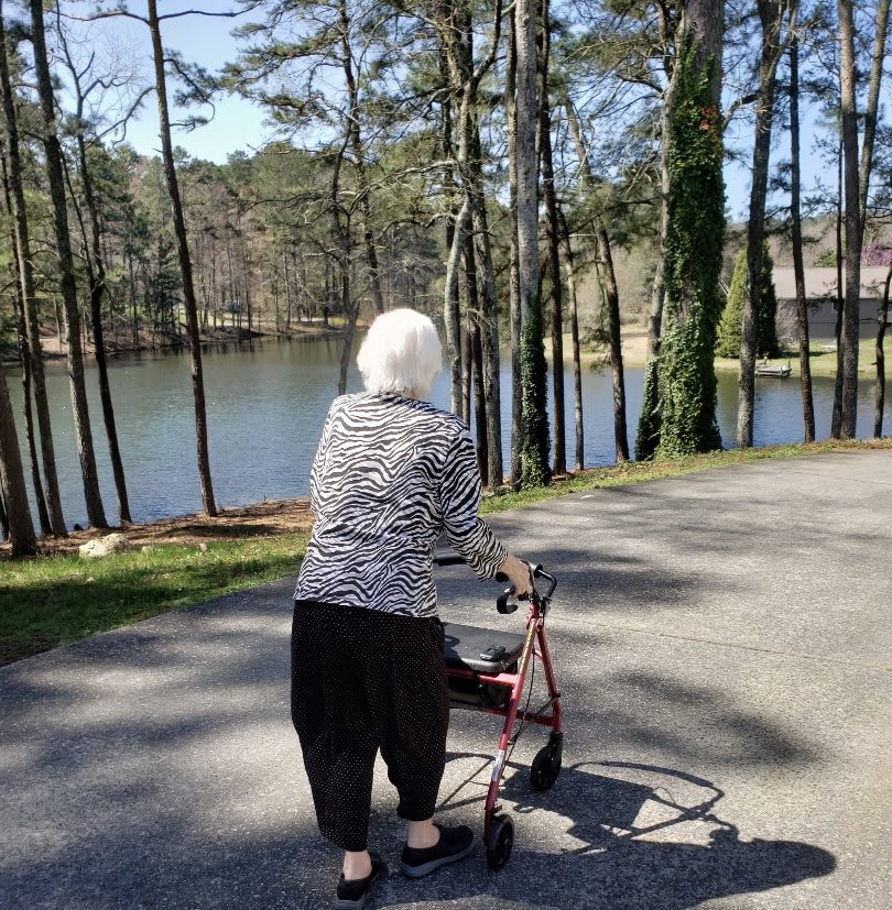 An elderly person using a walker near a lake surrounded by trees.