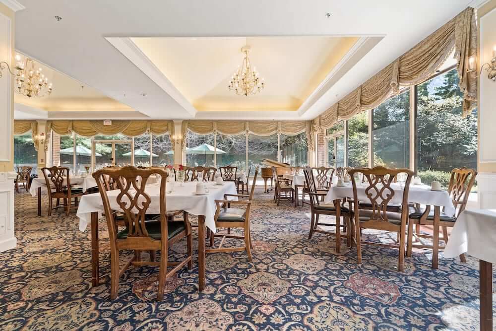 A spacious and elegant dining area with large windows allowing natural light to fill the room, featuring a chandelier and classic wooden furniture.