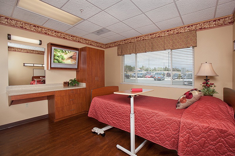 A hospital room featuring a single bed with red covers, a window, and medical furniture.