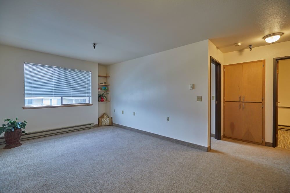 An empty room in an apartment with carpet flooring, a large window, and a doorway.