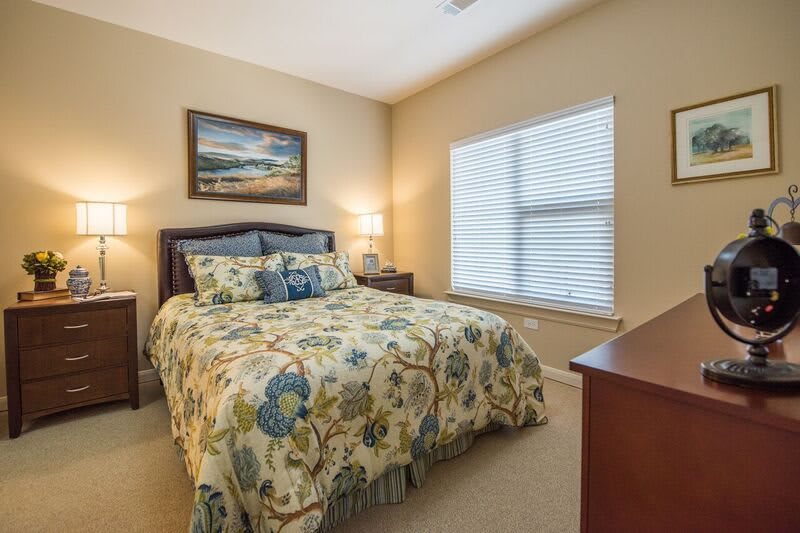 A cozy bedroom with floral bedding, featuring a window with blinds and a bedside lamp.