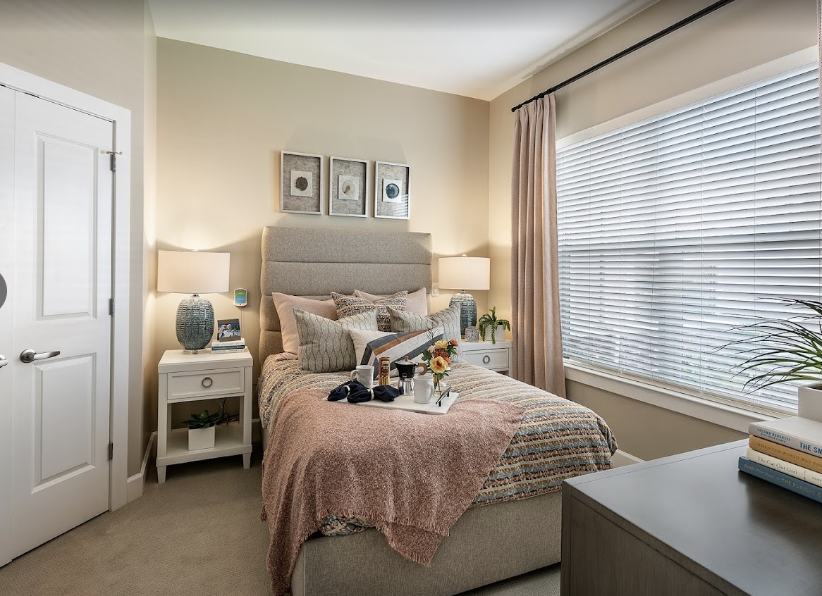A cozy bedroom with a neatly made bed, side tables with lamps, and large windows covered by curtains.