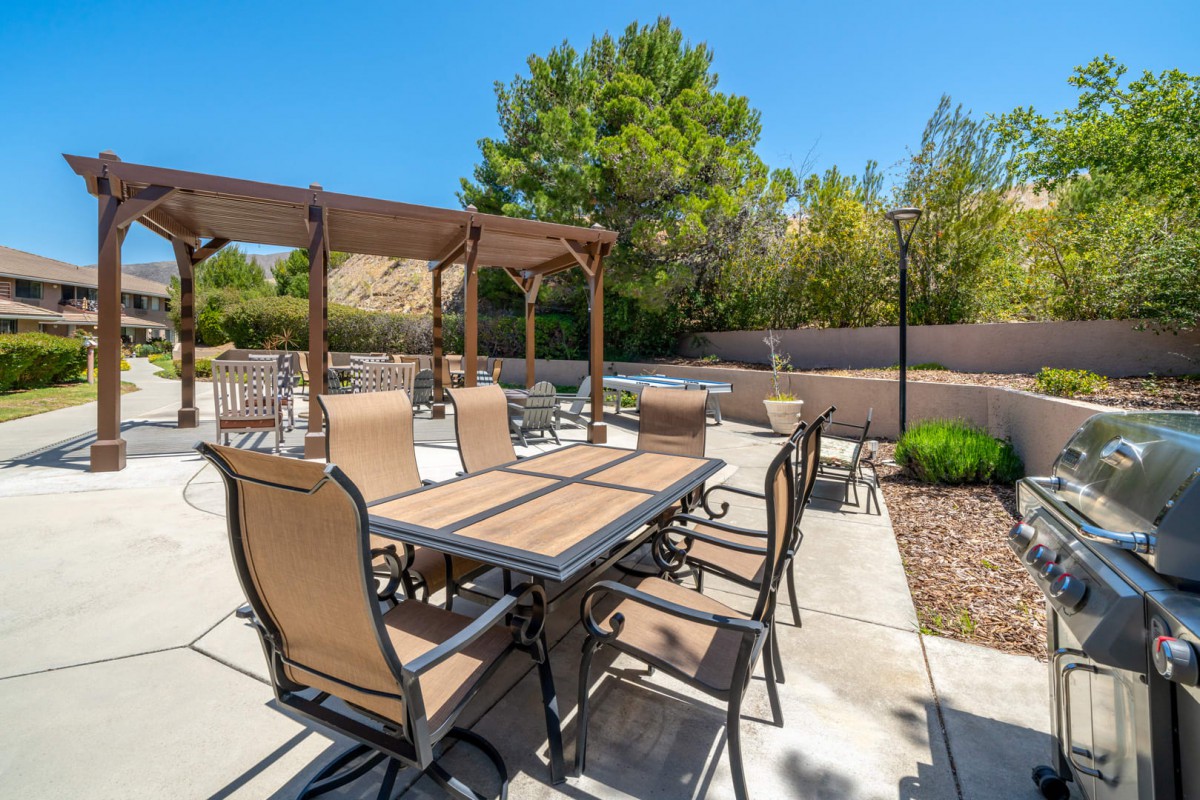 A sunny outdoor patio featuring a dining table with chairs, shaded by a pergola, surrounded by landscaped greenery.