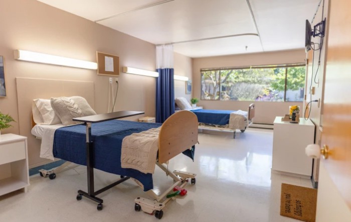 A hospital room with two beds, medical equipment, and a large window offering natural light.