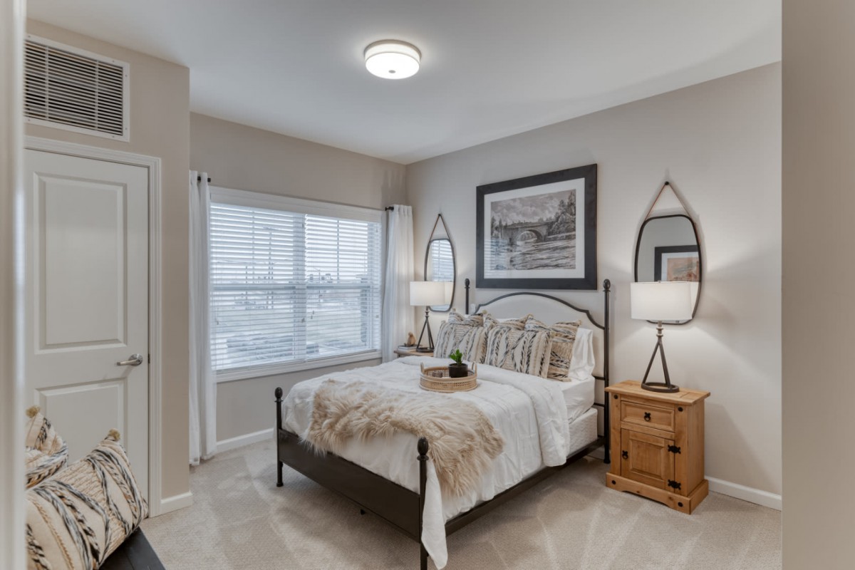 A cozy bedroom with neutral tones, featuring a neatly made bed, side tables with lamps, and a large window allowing natural light.